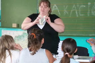 Illusztráció: tanárnő a zöld tábla előtt, kezében könyvvel, figyelő gyermekek fejei az előtérben